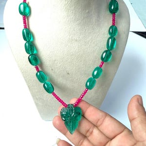 May include: A necklace featuring a central carved green leaf-shaped pendant, accented by alternating smooth oval green beads and small faceted pink beads. The strand connects to white seed beads at the top with gold-toned metal hardware connectors.