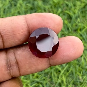 May include: A deep red gemstone with a round, faceted cut. The gemstone is held in a hand, with green grass visible in the background.