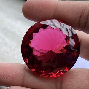 May include: A large, round, faceted gemstone with a deep red colour. The gemstone is cut in a brilliant style, with many facets that reflect light.