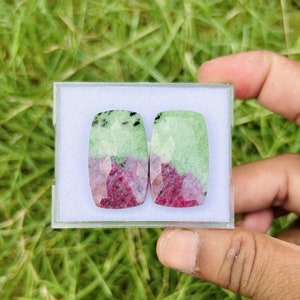 May include: Two rectangular gemstones with a faceted surface are displayed in a clear plastic case. The stones are divided into two colours: a vibrant red at the bottom and a light green at the top, with black inclusions.