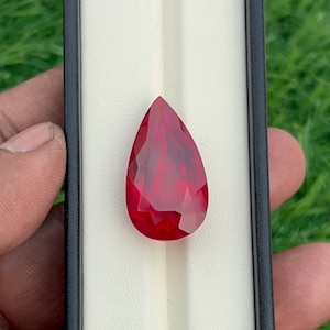 Pode incluir: Uma pedra preciosa vermelha escura facetada em forma de pêra. A gema é exibida em uma caixa preta com interior branco, em um fundo verde desfocado. O corte da pedra revela múltiplas facetas, realçando seu brilho.