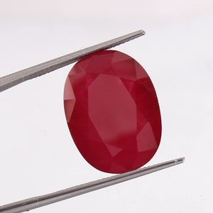 Puede incluir: Una piedra preciosa ovalada de color rojo oscuro sostenida por unas pinzas. La piedra preciosa está facetada y tiene una superficie lisa y pulida.