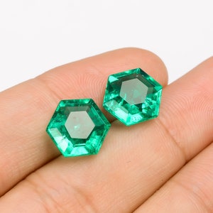 Puede incluir: Dos piedras preciosas de color verde esmeralda, talladas en forma hexagonal, con un diseño facetado. Las piedras preciosas se muestran en una mano.