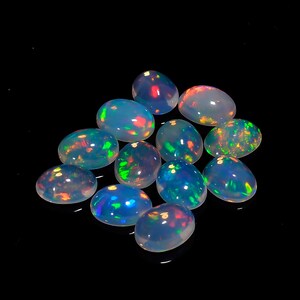 May include: A group of 12 oval-shaped opal gemstones with iridescent colors, including blue, green, red, and orange. The opals are arranged in a loose pile on a black background.