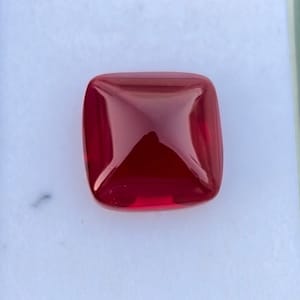 Pu&ograve; includere: Una pietra preziosa di colore rosso intenso con forma quadrata e parte superiore leggermente bombata.