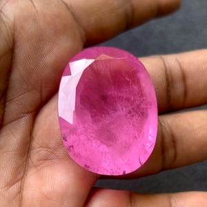 May include: A large, oval-cut gemstone in a vivid pink colour. The gem has a polished surface with multiple facets that reflect light, creating a sparkling effect. The stone is held in a hand, showing its size and colour.