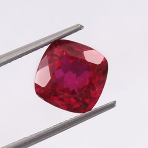 May include: A deep red gemstone with a cushion cut. The gemstone is held in tweezers against a white background.