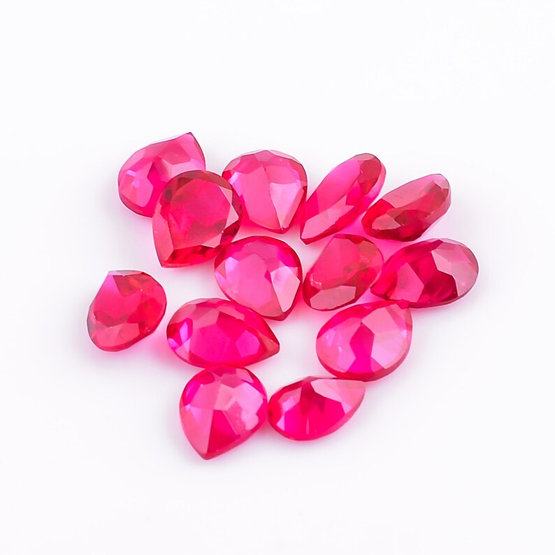 May include: A collection of faceted, teardrop-shaped, hot pink gemstones. The gems are scattered on a white surface, each catching the light with multiple facets. The stones vary slightly in size and shape, creating a dynamic display.
