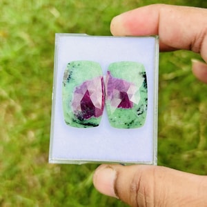 May include: Two faceted ruby in zoisite gemstones, each with a rectangular shape and rounded corners. The stones feature a vibrant mix of green, black, and deep red hues, displayed in a clear plastic case.