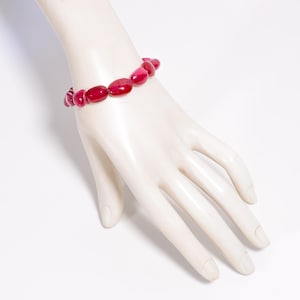 Op de afbeelding: Een opvallende rode kralenarmband. De armband bestaat uit ovaalvormige kralen, geregen tot een opvallend accessoire. De kralen hebben een glanzende afwerking die het licht reflecteert. De armband wordt getoond op een witte mannequinhand.