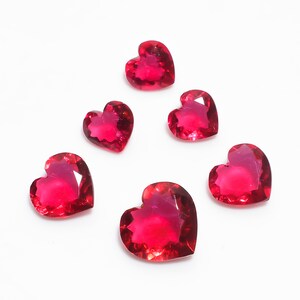 May include: Five heart-shaped red gemstones on a white background. The gemstones are faceted and have a shiny finish.