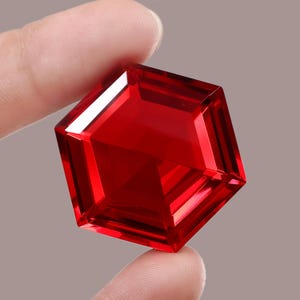 May include: A square-cut, deep red gemstone with multiple facets. The gem is held between two fingers, highlighting its reflective surfaces. The gem's colour is a vibrant, saturated red, with sharp edges and a polished finish.