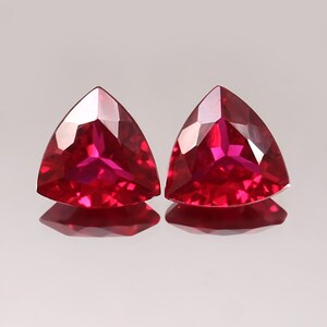 Puede incluir: Dos gemas triangulares facetadas de color rojo intenso. Las gemas se muestran sobre una superficie reflectante, mostrando sus brillantes facetas y color. Probablemente sean rubíes, comúnmente utilizados en joyería.