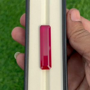 May include: A vibrant red gemstone with a rectangular cut, displayed in a white velvet box with a black border.