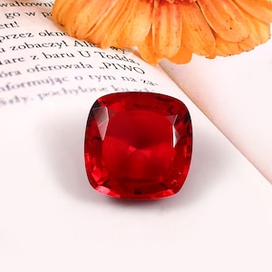 Peut inclure: Une pierre précieuse rouge vif, taillée en coussin, avec de multiples facettes. La gemme est posée sur un fond blanc avec une fleur orange et du texte d'un livre partiellement visibles.