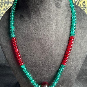 May include: A beaded necklace featuring alternating sections of translucent emerald green and ruby red faceted glass beads. The strand is punctuated by a single, larger, smooth dark red glass bead positioned at the center of the necklace.