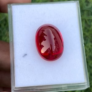 Op de afbeelding: Een helderrode ovale edelsteen in een witte doos. De edelsteen is glad en glanzend.