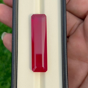 May include: A deep red rectangular gemstone with a faceted cut, displayed in a white box with a black border.