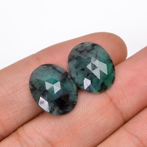 Könnte beinhalten: Zwei ovale, grüne Smaragde mit schwarzen Einschlüssen. Die Smaragde sind facettiert geschliffen und in einer Zargenfassung gefasst.