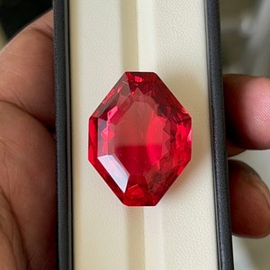 May include: A deep red octagonal gemstone with a faceted cut. The gemstone is displayed on a white background.
