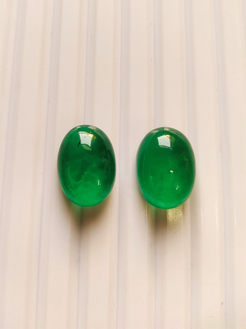 Puede incluir: Dos piedras preciosas verdes de forma ovalada, probablemente esmeraldas, se muestran sobre una superficie blanca. Las piedras preciosas son lisas y pulidas, reflejando la luz.