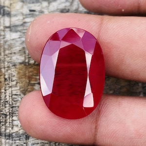 Pode incluir: Uma pedra preciosa de rubi facetada em forma oval. A gema é de uma cor vermelha profunda e saturada com um alto grau de clareza. O corte da pedra cria múltiplas superfícies refletoras, realçando o seu brilho.
