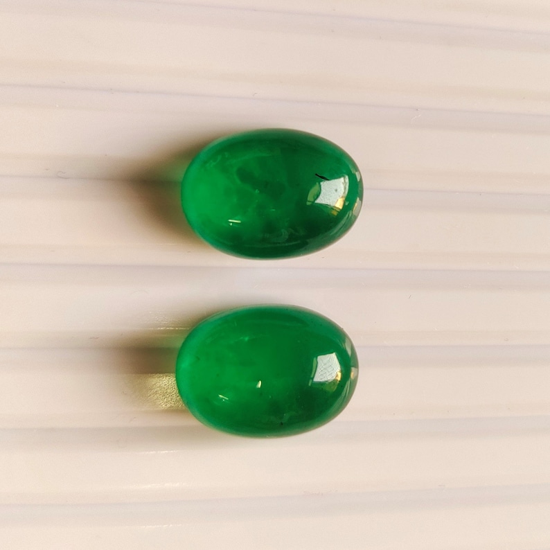 Puede incluir: Dos piedras preciosas verdes de forma ovalada sobre un fondo blanco.