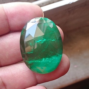 May include: A large, oval-shaped, faceted emerald gemstone with a deep green colour. The stone has a slightly cloudy appearance, with visible inclusions.