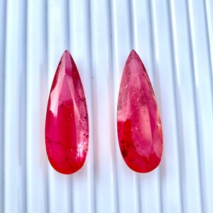 May include: Two elongated, faceted gemstones with a vibrant red hue. The gems have a teardrop shape, tapering to a point at one end. The stones are displayed against a white, ridged surface.