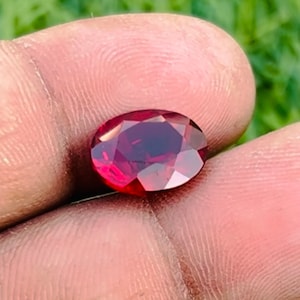 May include: A faceted, oval-shaped, deep red gemstone. The gem is held between fingers, with the green grass in the background. The gem has a polished, reflective surface, showcasing its internal structure and color.