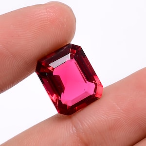 Puede incluir: Una gema roja vibrante, de corte esmeralda rectangular. La gema se sostiene entre dos dedos, mostrando su brillo y claridad.