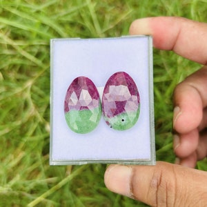 Może przedstawiać: Dwa owalne kamienie szlachetne, jeden zielony, a drugi różowy, o fasetowanym szlifie. Kamienie szlachetne są prezentowane w białym pudełku.