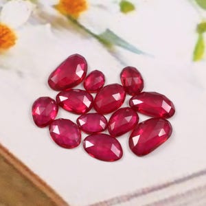 Puede incluir: Una colección de piedras preciosas rojas profundas, facetadas y de forma ovalada. Las piedras están dispuestas sobre una superficie blanca, mostrando sus superficies pulidas y facetas internas. Los tamaños y formas variados de las piedras crean interés visual.