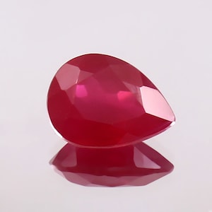 Puede incluir: Una piedra preciosa facetada en forma de pera, de color rojo intenso. La piedra preciosa está colocada sobre un fondo blanco.