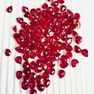 May include: A large group of pear-shaped ruby gemstones, all of a deep red colour, are scattered on a white surface.