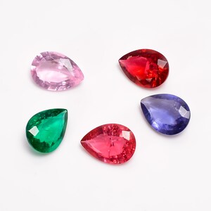 Puede incluir: Cinco piedras preciosas en forma de pera de diferentes colores: rosa, rojo, verde, azul y otro tono de rojo. Las piedras preciosas están dispuestas en semicírculo sobre un fondo blanco.