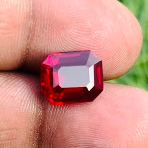 May include: A deep red gemstone with a rectangular emerald cut. The gemstone is faceted and has a high polish.