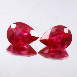 Puede incluir: Dos piedras preciosas rojas en forma de pera con un corte facetado. Las piedras preciosas están colocadas sobre un fondo blanco.