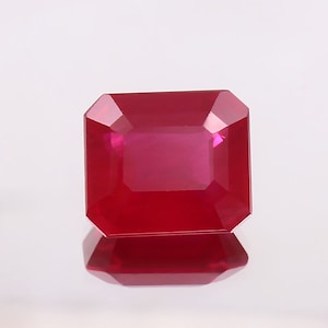 Könnte beinhalten: Ein leuchtend roter Edelstein mit quadratischem Smaragdschliff. Der Edelstein ist facettiert und hochglanzpoliert.