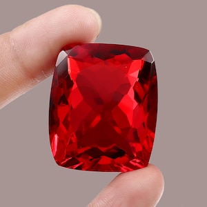 May include: A large, faceted, cushion-cut gemstone with a deep red hue. The gem is held between fingers, showcasing its brilliant, reflective surfaces. The gemstone's shape is rectangular with rounded corners, and it appears to be a ruby.