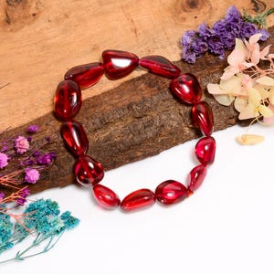 May include: A red gemstone bracelet with smooth, oval-shaped beads. The bracelet is displayed on a piece of wood with dried flowers.