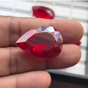 Puede incluir: Una piedra preciosa de color rojo intenso en forma de pera con un corte facetado. La piedra preciosa se sostiene en una mano.