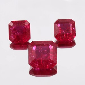 May include: Three faceted ruby gemstones, each with a rectangular shape and beveled edges. The gems exhibit a deep red colour and are arranged on a reflective surface, enhancing their brilliance. The gems are approximately 1.5 cm in length.