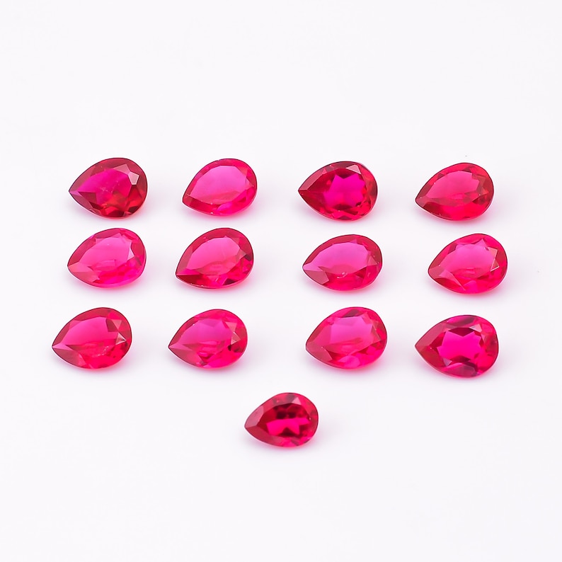 May include: A collection of fourteen faceted, teardrop-shaped gemstones. The gems are a vibrant, translucent red color, arranged on a white background. Each stone has a pointed base and a rounded top, catching the light to create a sparkling effect. Ideal for jewelry making.