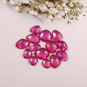Puede incluir: Una colección de gemas facetadas de color rosa intenso en varias formas, incluyendo redondas, ovaladas y en forma de lágrima. Las gemas están dispersas sobre una superficie blanca, con un arreglo floral borroso en el fondo.