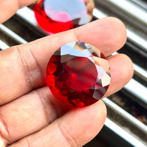 May include: A faceted, round, deep red gemstone is held in a hand. The gem has multiple reflective surfaces, catching light and creating a brilliant display. Another similar gem is visible in the background.
