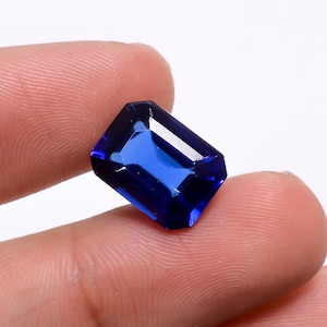 May include: A deep blue gemstone with a rectangular emerald cut. The gemstone is held in a hand.