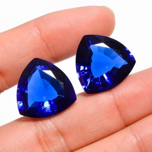 May include: Two blue triangular gemstones with a faceted cut. The gemstones are held in a hand.