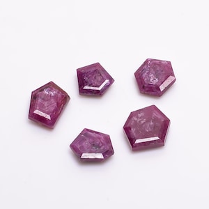 May include: Five faceted, pink ruby gemstones, each with a hexagonal shape, are arranged on a white surface.