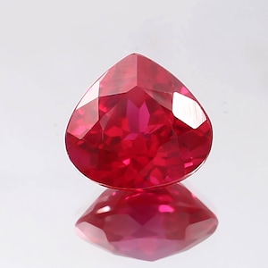 May include: A vibrant red pear-shaped gemstone with a faceted cut. The gemstone is set against a white background and reflects light.